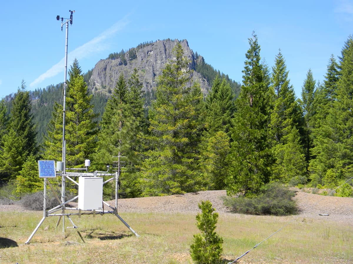Solar-powered remote monitoring station with equipment enclosure and mast beside a rocky forest clearing